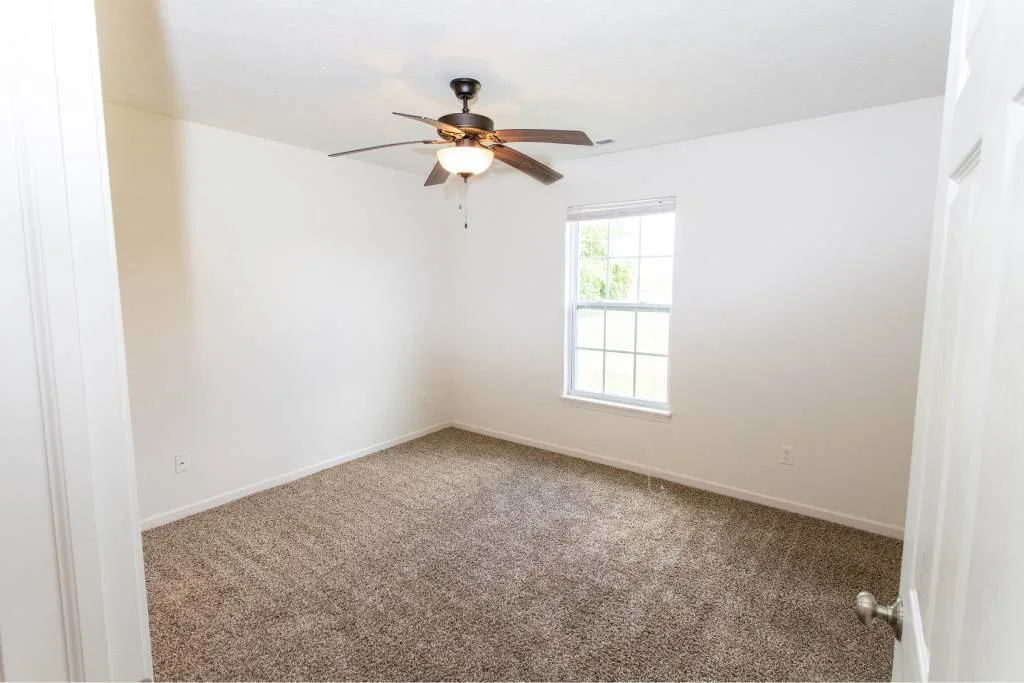 Empty room with white walls, carpeted floor, ceiling fan with light, and a window letting in natural light.