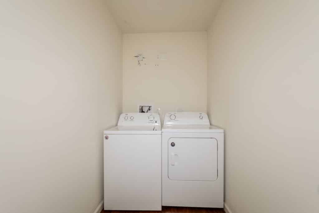 A washing machine and dryer are positioned side by side in a small, empty laundry room with beige walls and a wooden floor, showcasing the functional design found throughout Gallery - McCormick Manor.