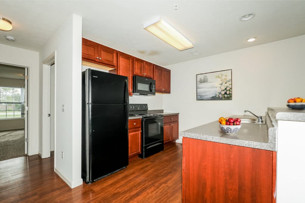 Modern apartment kitchen at Gallery - McCormick Manor with dark wood cabinets, black appliances, gray countertops, and a bowl of fruit on the counter; open view to a bedroom with carpeted floor.
